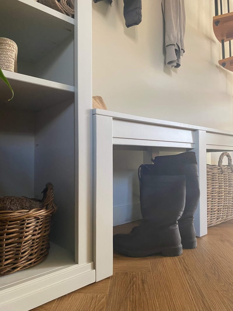 Low view of the join between the shelf unit and the bench, showing the plinth and open bays with a wicker basket, and tall black boots and a handled basket tucked beneath the bench