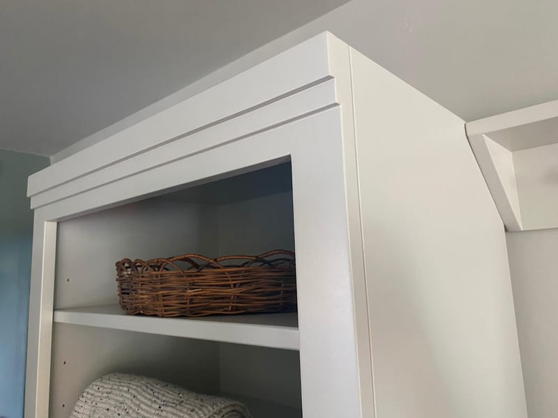 Close view of the top corner of the white shelf unit, showing the stepped cornice, the adjustable shelf peg holes and a shallow wicker tray on the top shelf, with the peg rail beyond