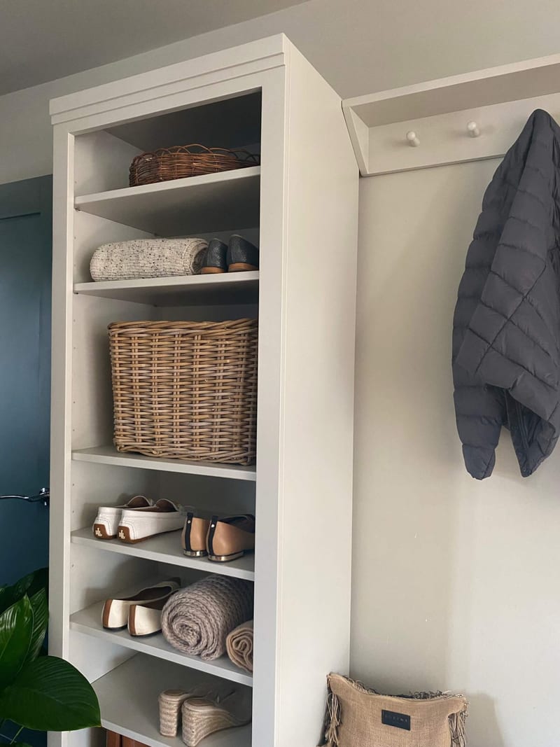 View of the tall open shelf unit from the front and side, its shelves holding baskets, loafers, heeled mules, espadrilles and rolled throws, beside the peg rail and a charcoal quilted jacket