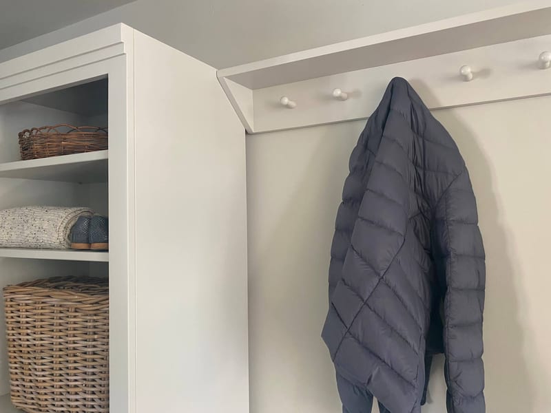 Close view where the peg rail meets the side of the shelf unit, showing turned white pegs, a charcoal quilted jacket hanging and the cornice, wicker tray and kubu basket to the left