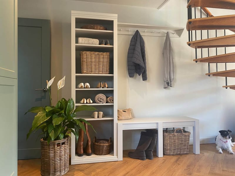 Landscape view of the whole boot room run, with the shelf unit, peg rail, two coats and twin benches, plus a peace lily in a basket and a Jack Russell terrier on the oak floor