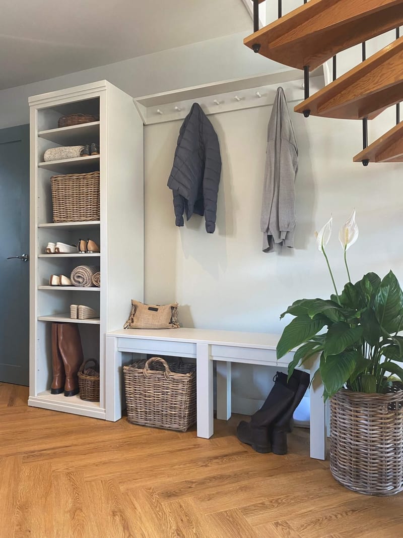 Angled view of the boot room from the side, showing the shelf unit with tan boots at its base, the peg rail below a wooden spiral staircase and two benches with a wicker basket beneath