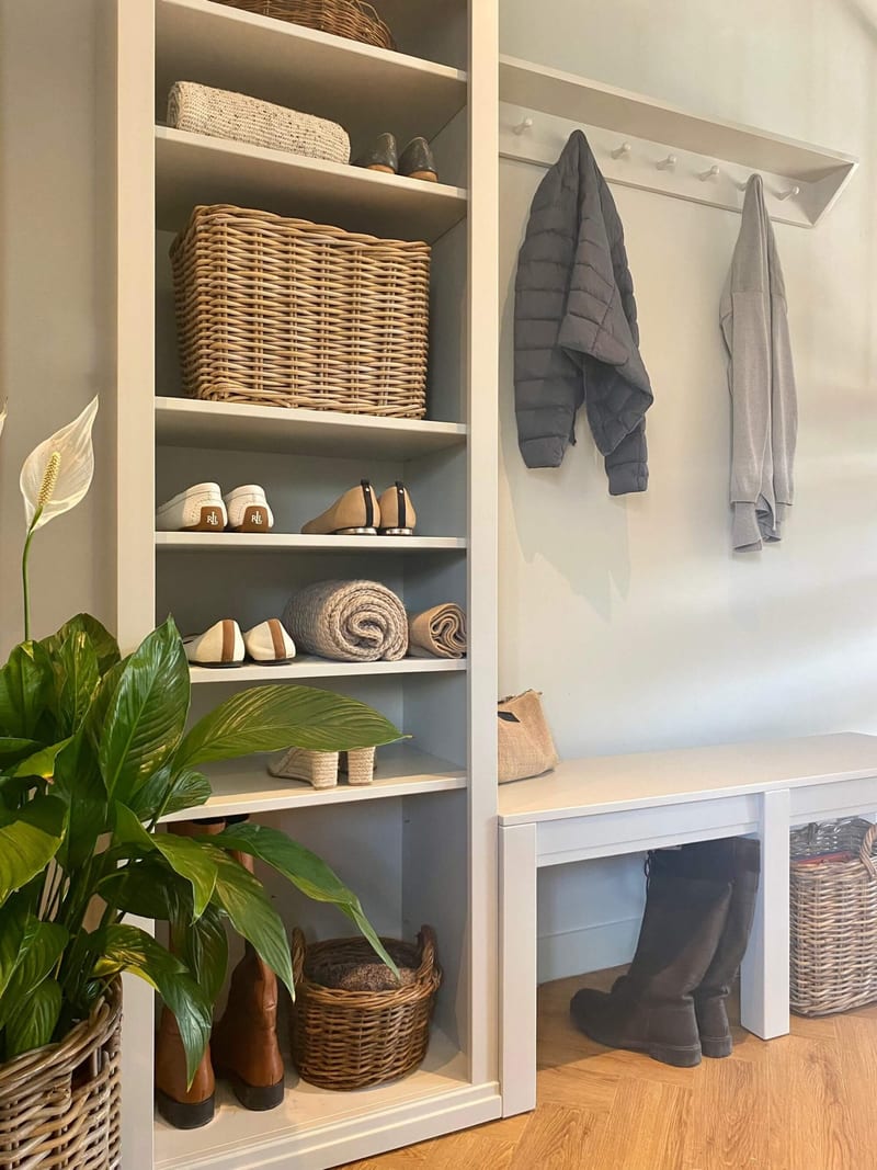 Three-quarter view of the shelf unit and peg rail together, with a folded knit, a wicker basket, shoes and rolled throws on the shelves, and a peace lily leaning in from the left