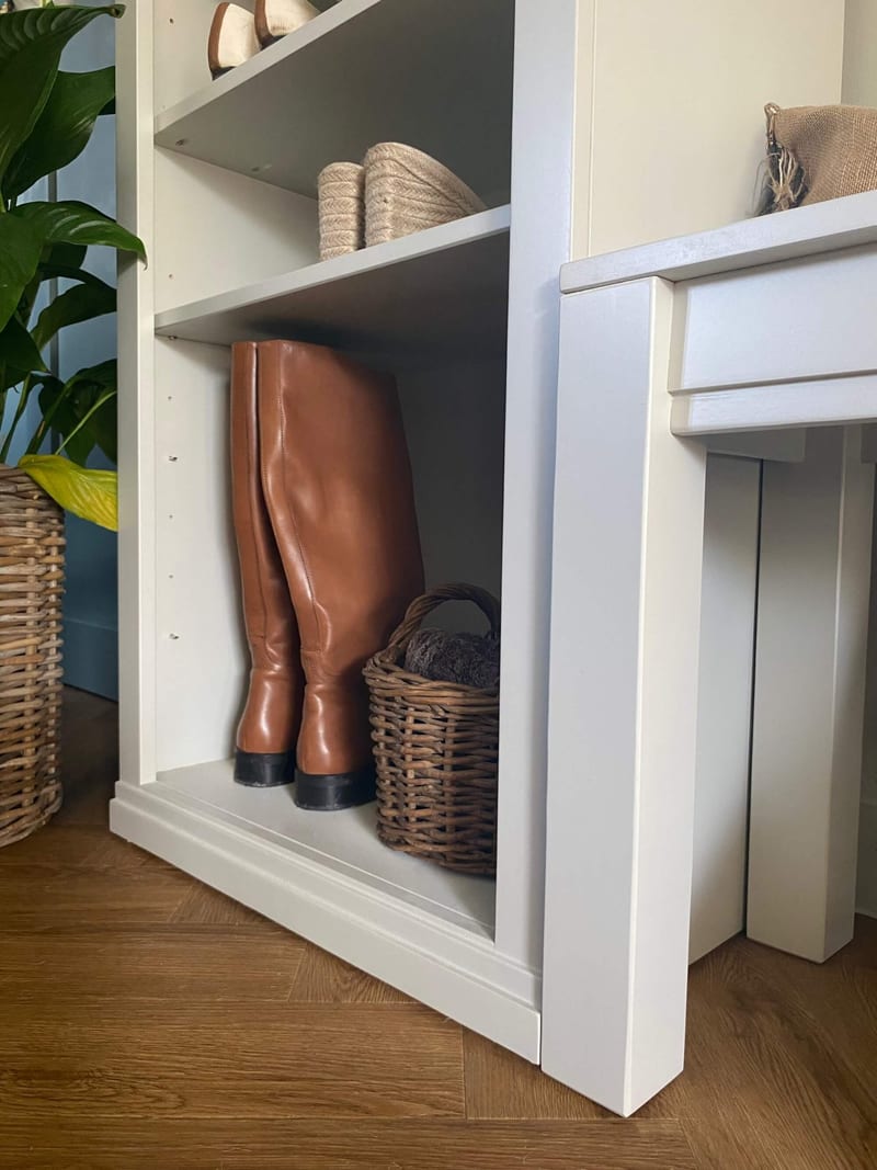 Close view of the open bottom bay of the shelf unit, holding tan leather knee boots and a small round wicker basket above the moulded plinth, beside the painted frame of the bench