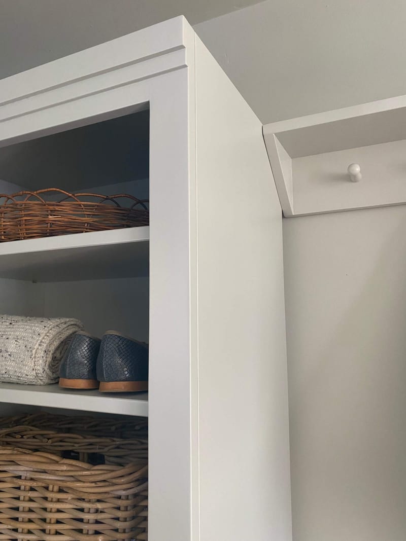 Close view of the top corner and side panel of the shelf unit, showing the moulded cornice, a wicker tray, a folded knit and dark blue loafers, with a white peg on the rail behind