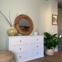 White painted Colmworth double chest of six drawers, beneath a round rattan mirror with a gold vase of pussy willow, beside a jute pouffe and a peace lily in a seagrass basket