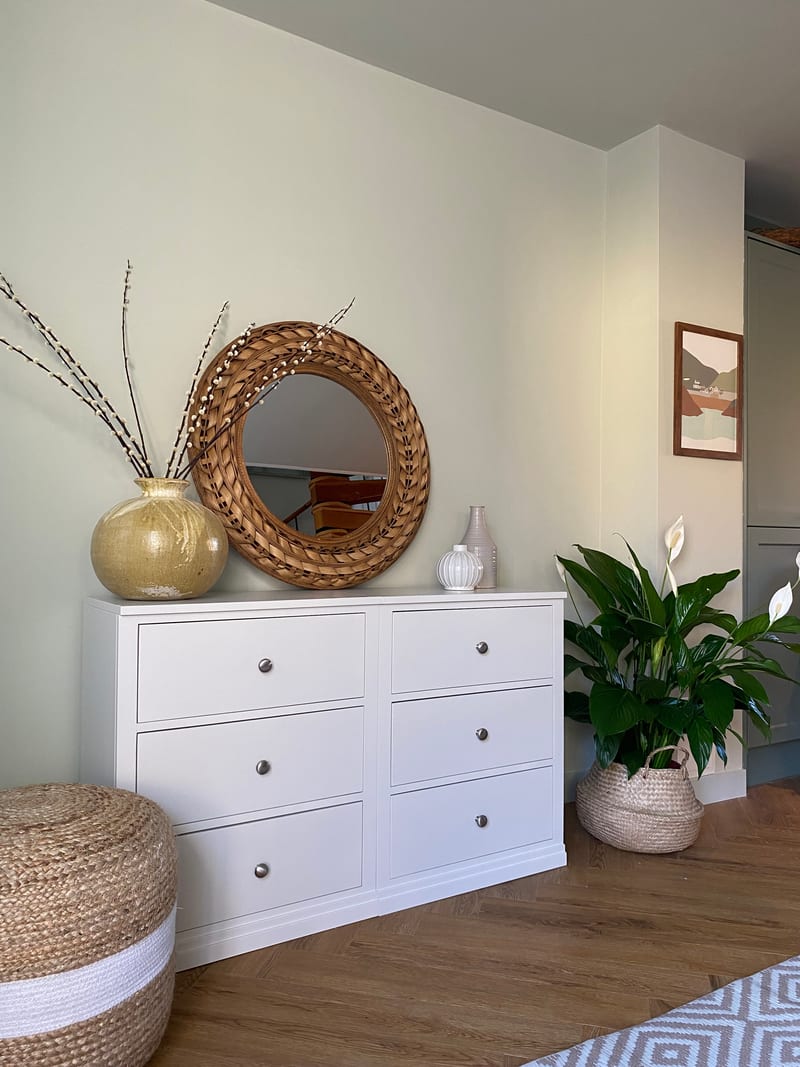 White painted Colmworth double chest of six drawers, beneath a round rattan mirror with a gold vase of pussy willow, beside a jute pouffe and a peace lily in a seagrass basket