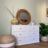 Straight-on view of the white six-drawer double chest, two banks of three drawers with brushed nickel knobs, topped by a round rattan mirror, a gold vase of catkins and two pale vases