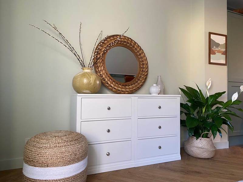 Straight-on view of the white six-drawer double chest, two banks of three drawers with brushed nickel knobs, topped by a round rattan mirror, a gold vase of catkins and two pale vases