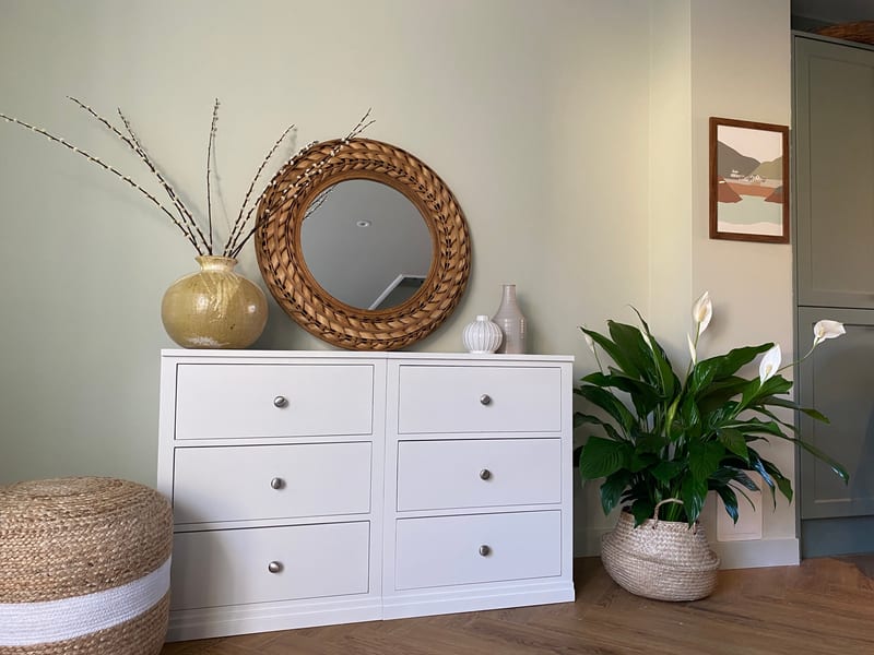 Wide view of the six-drawer chest against a pale sage wall, with the rattan mirror, a gold glazed vase of pussy willow, a peace lily in a basket and a framed coastal print alongside
