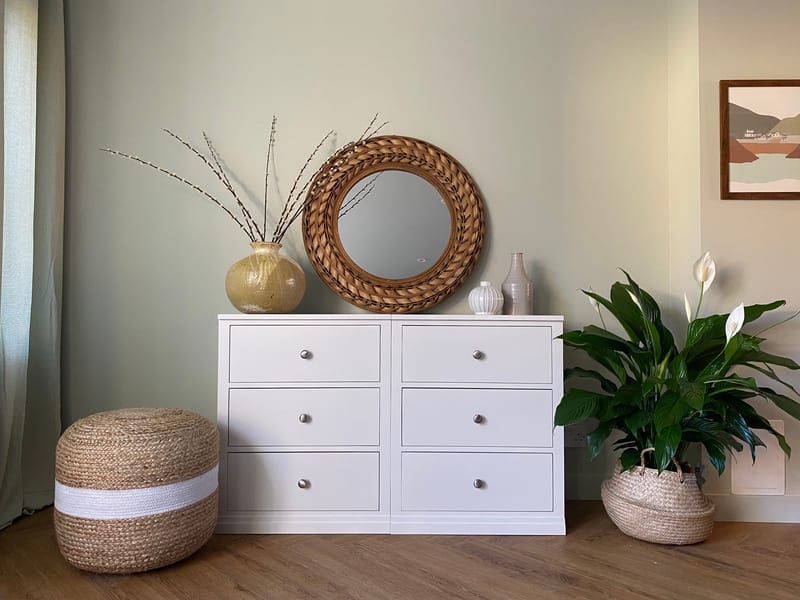 Full landscape view of the white double chest of drawers on oak flooring, flanked by a striped jute pouffe and a peace lily in a basket, beneath a round woven rattan mirror