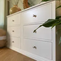 Low three-quarter view of the chest from the right, showing six drawers with brushed nickel knobs, the stepped plinth and a peace lily leaf crossing the frame