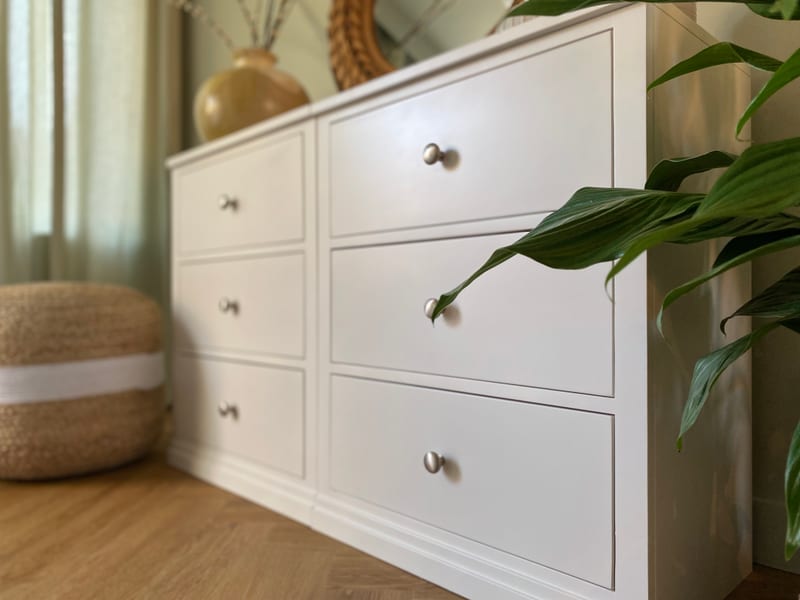 Low three-quarter view of the chest from the right, showing six drawers with brushed nickel knobs, the stepped plinth and a peace lily leaf crossing the frame