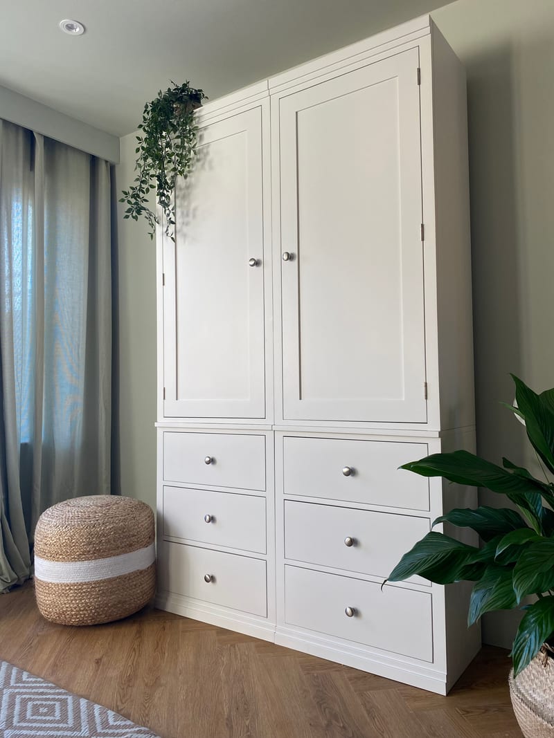 White painted Colmworth modular double wardrobe with two tall panelled doors above six drawers, with trailing ivy on top and a jute pouffe alongside