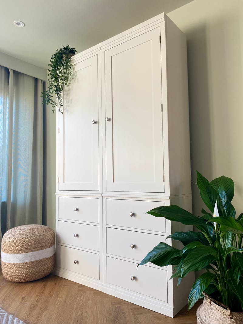 Angled view of the white painted double wardrobe showing two panelled doors above six drawers with brushed nickel knobs, a trailing faux plant on top and a jute pouffe on the oak floor