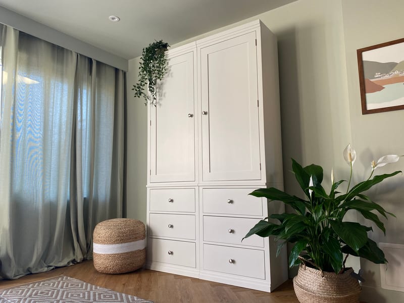 Wider landscape view of the white painted double wardrobe in a sage green bedroom, with linen curtains, a striped jute pouffe, a peace lily in a seagrass basket and a framed coastal print