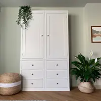 Straight-on view of the white painted double wardrobe, two panelled doors above six drawers, with a jute pouffe to the left and a peace lily, framed coastal print and sage green cupboard to the right