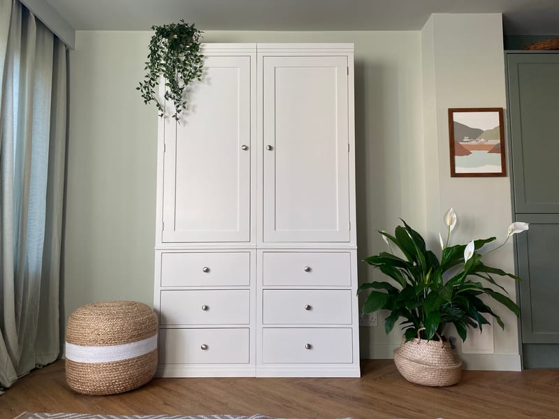 Straight-on view of the white painted double wardrobe, two panelled doors above six drawers, with a jute pouffe to the left and a peace lily, framed coastal print and sage green cupboard to the right