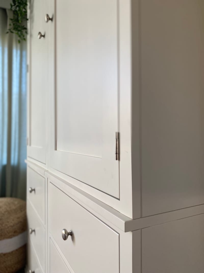 Side angle detail of the white painted wardrobe, showing the plain painted end panel, a metal butt hinge on the door edge and round brushed nickel knobs on the drawers below