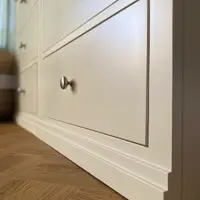 Low close view of the drawer fronts and moulded plinth of the white painted wardrobe base, with round brushed nickel knobs on both banks of drawers and herringbone oak flooring beneath