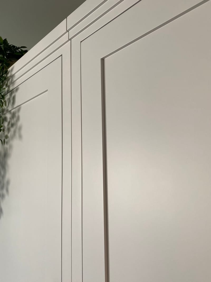 Close view of the top corner of the white painted wardrobe, showing the stepped cornice moulding, the panelled door fronts and the shadow of a trailing plant cast across the paintwork