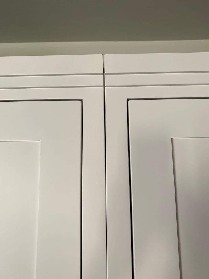 Detail of the tops of the two white painted wardrobe doors, showing the stepped cornice beading, the panel mouldings and the narrow gap where the doors meet below the shadowed ceiling