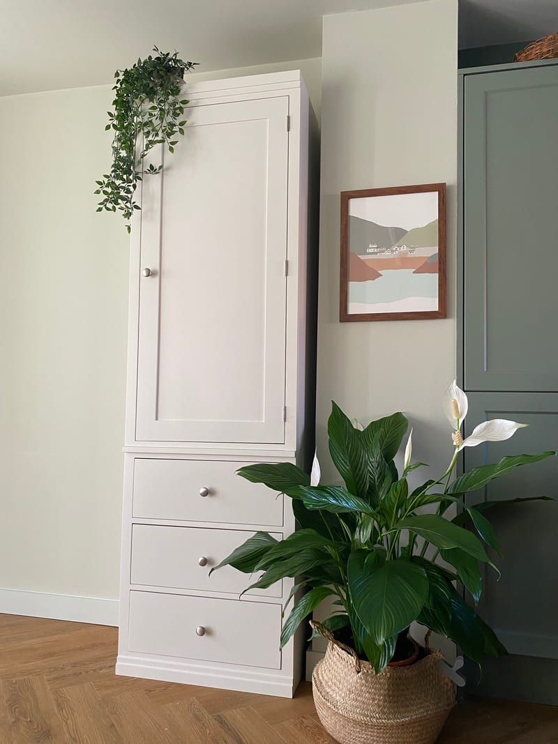 White painted Colmworth modular wardrobe with a single tall panelled door above three drawers, with trailing ivy on top and a peace lily in a seagrass basket alongside