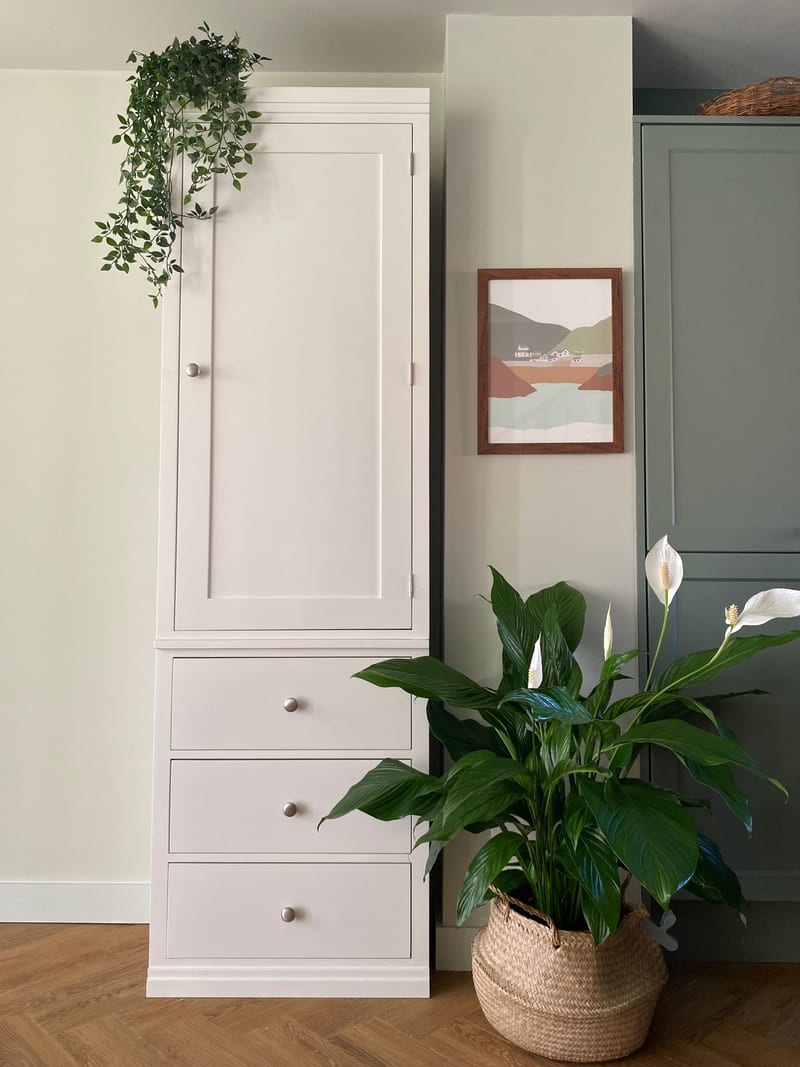 Front view of the tall narrow white painted wardrobe, one panelled door above three drawers with brushed nickel knobs, beside a peace lily and a framed coastal print on a pale green wall