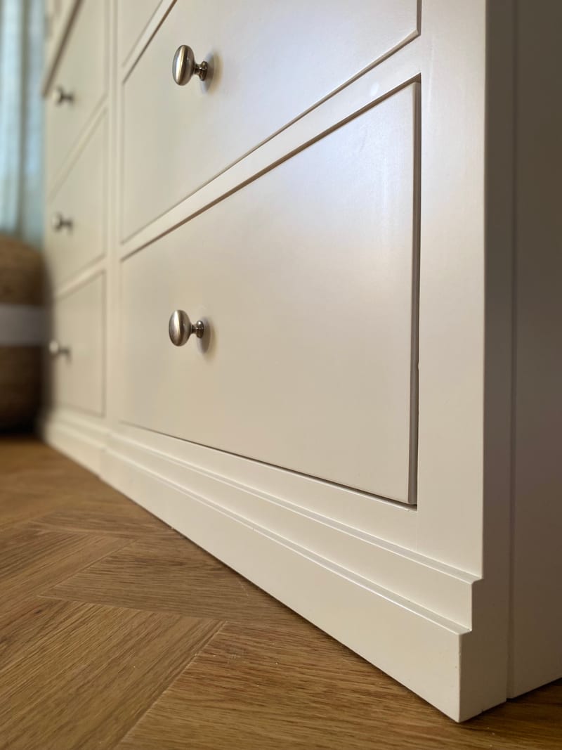 Low close view of the white painted wardrobe drawer fronts, showing round brushed nickel knobs, the fine moulded drawer edges and the shaped plinth above herringbone oak flooring