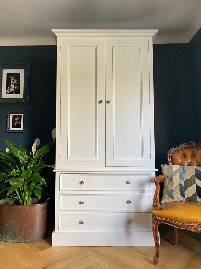 Cream painted double wardrobe with two tall panelled doors above three deep drawers, against a dark navy wall with a peace lily in a copper pot and a tan leather chair alongside