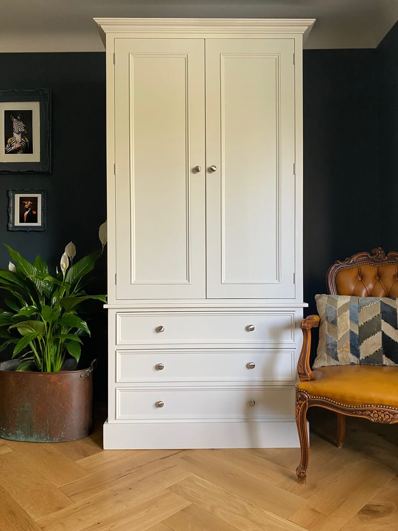 Cream painted double wardrobe with two tall panelled doors above three deep drawers, against a dark navy wall with a peace lily in a copper pot and a tan leather chair alongside