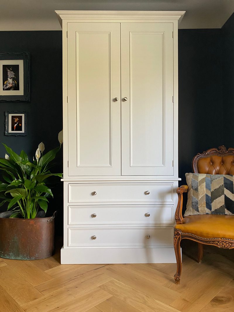 Front view of the cream painted double wardrobe against a near black wall, showing the moulded cornice, two panelled doors and three wide drawers with brushed nickel knobs, on herringbone oak flooring