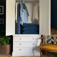 Both doors of the cream painted wardrobe open to show the hanging rail with shirts on white hangers, a tongue-and-groove back panel and folded blue cushions stacked on the drawer top