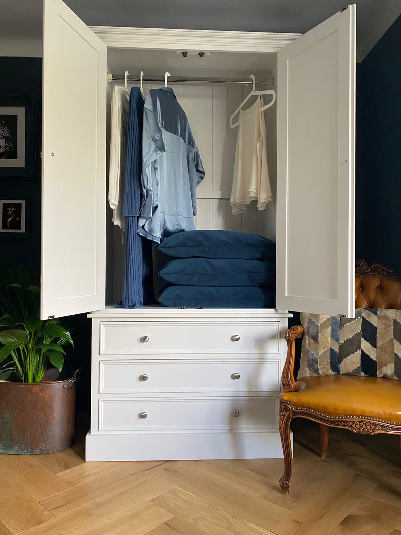 Both doors of the cream painted wardrobe open to show the hanging rail with shirts on white hangers, a tongue-and-groove back panel and folded blue cushions stacked on the drawer top