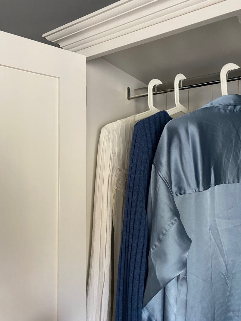 Close view inside the open wardrobe, showing the chrome hanging rail on its bracket, white hangers and a cream blouse, a navy knit and a blue satin shirt hanging below the moulded cornice