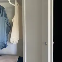 Detail of the open wardrobe door edge with its small chrome knob, framing a cream camisole and a blue satin shirt hanging inside against the pale painted interior