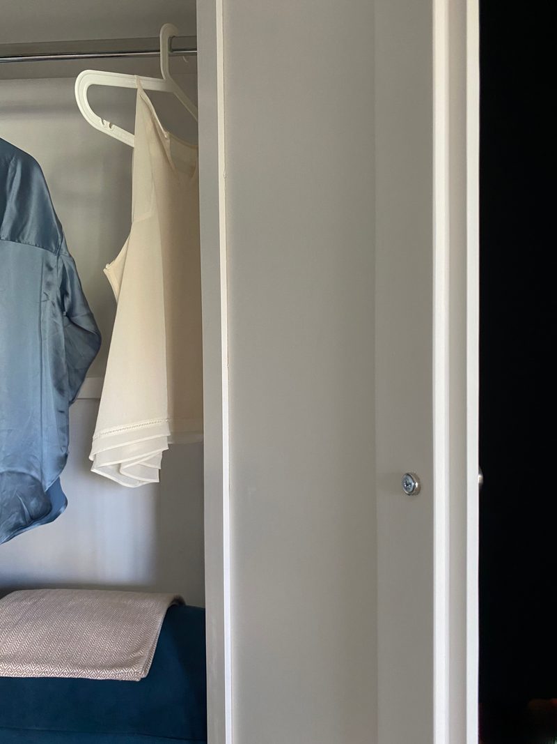 Detail of the open wardrobe door edge with its small chrome knob, framing a cream camisole and a blue satin shirt hanging inside against the pale painted interior