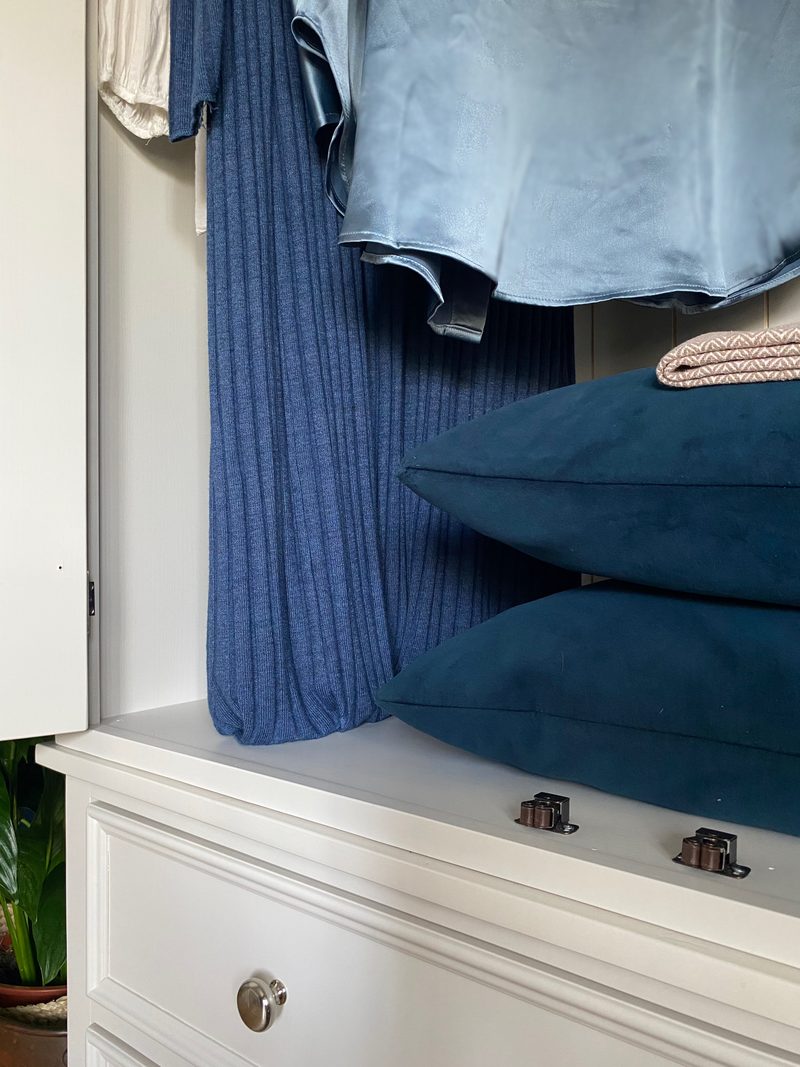 Close view inside the wardrobe, showing stacked teal cushions and a folded throw resting on the painted top of the drawer base beside two dark metal fixing brackets