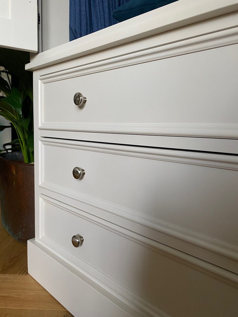Angled close view of the three drawer fronts of the cream painted wardrobe, showing the moulded edges, round brushed nickel knobs and the moulded plinth above oak flooring