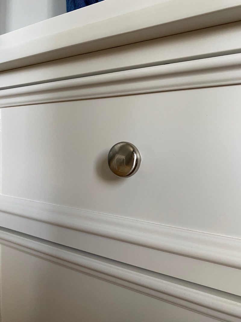 Extreme close-up of a single round brushed nickel knob on a cream painted drawer front, showing the fine moulded beading running above and below the drawer face