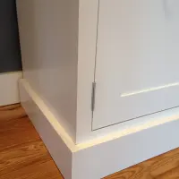 Close view of the corner of the white painted sideboard at floor level, showing the deep plinth base, a steel butt hinge on the shaker door and the oak floorboards beneath