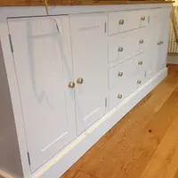 Low angled view along a white painted dresser base, showing the shaker doors, four drawers with brushed nickel knobs, the waxed pine top with a plant and candlestick and the boarded panel behind