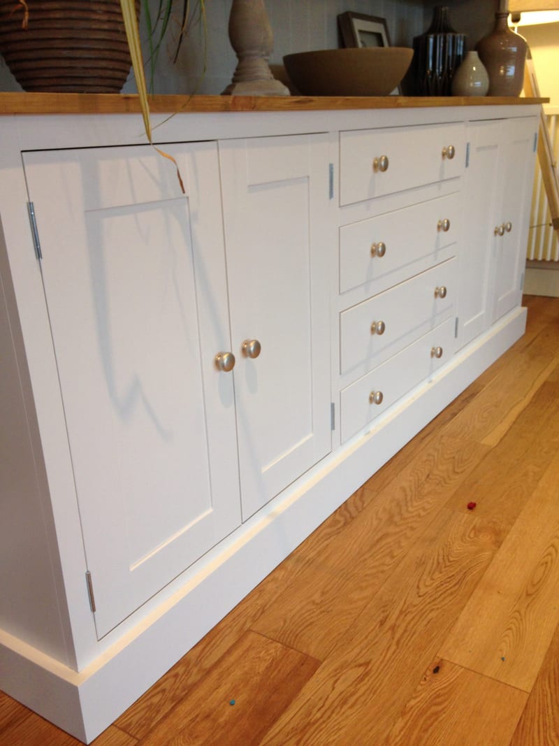 Low angled view along a white painted dresser base, showing the shaker doors, four drawers with brushed nickel knobs, the waxed pine top with a plant and candlestick and the boarded panel behind