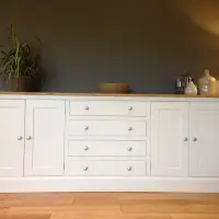 Full front view of the white painted sideboard against a dark grey wall, with four shaker doors, four drawers, brushed nickel knobs and a pine top holding a plant, bowl and vases