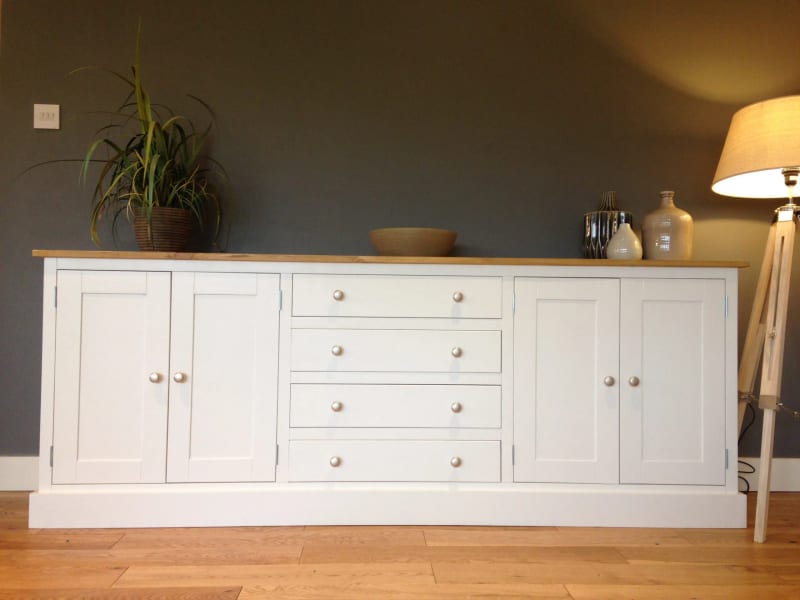 Full front view of the white painted sideboard against a dark grey wall, with four shaker doors, four drawers, brushed nickel knobs and a pine top holding a plant, bowl and vases