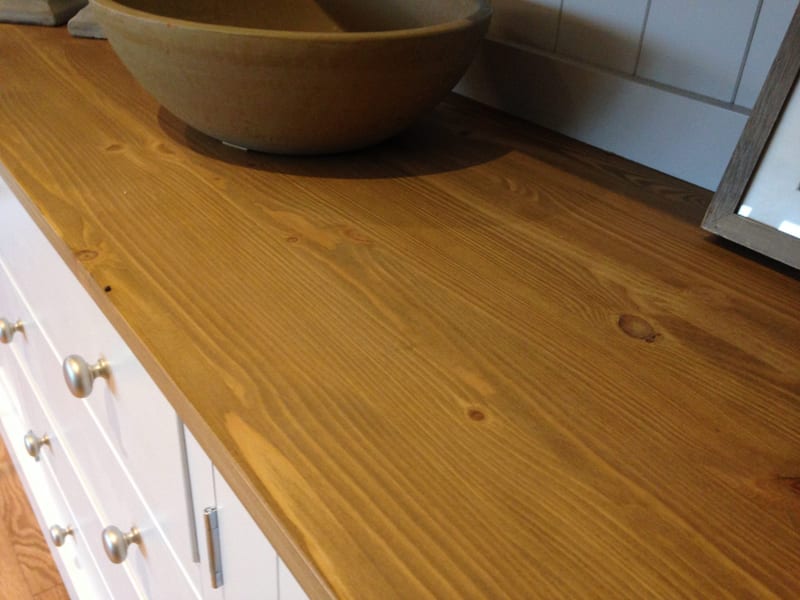 Close view along the waxed pine worktop of the dresser, showing the grain and knots beside a stoneware bowl, with white painted drawer fronts, brushed nickel knobs and a steel butt hinge below
