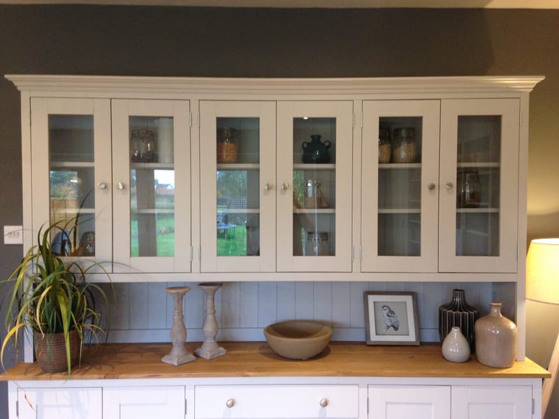 Front view of the glazed upper rack, showing the moulded cornice, six glass doors with brushed nickel knobs, storage jars on the shelves and the pine top below holding candlesticks and a bowl