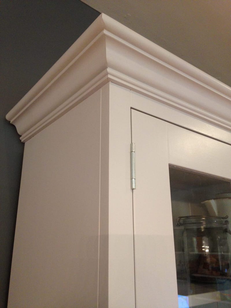 Close view of the top corner of the painted rack, showing the layered moulded cornice, a steel butt hinge on the glazed door and a storage jar behind the glass