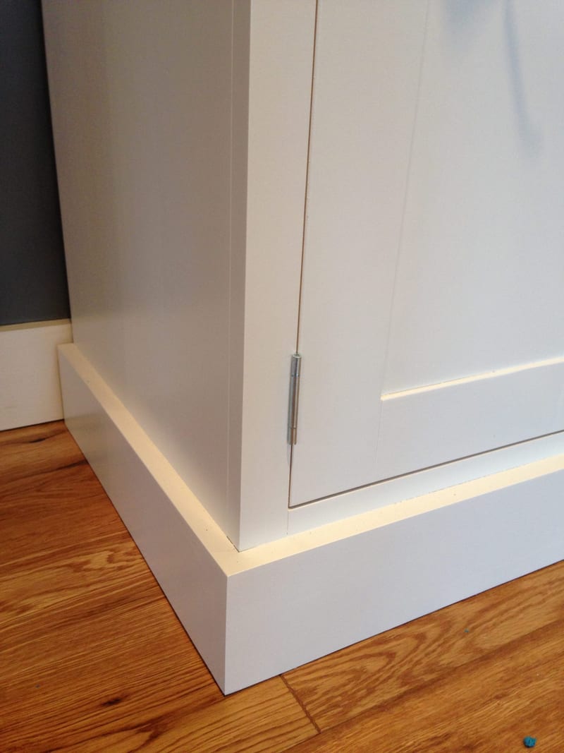 Detail of the base corner at floor level, showing the stepped plinth, the painted door edge with its steel hinge and the oak boards and white skirting behind