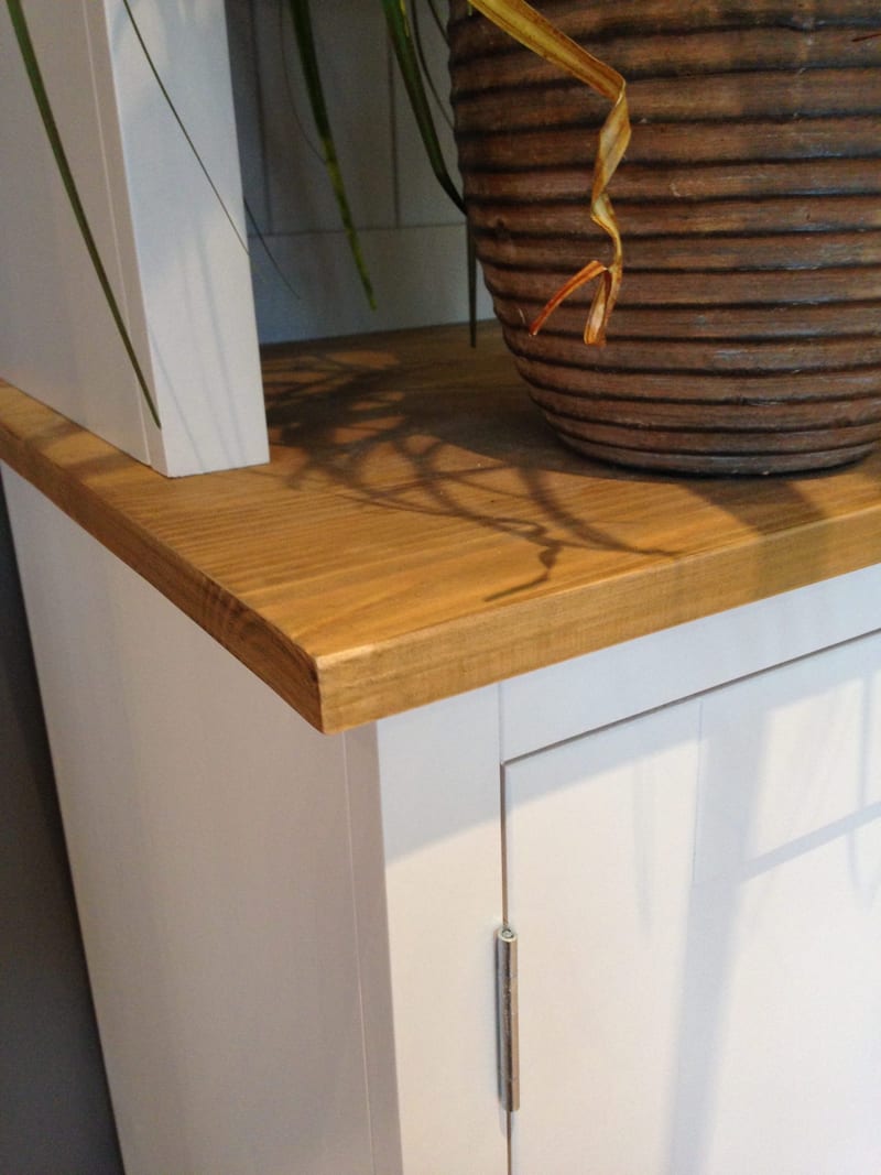 Close-up of the corner of the pine worktop, showing the grain and squared edge beside a ribbed terracotta plant pot, with the white painted side and a steel door hinge below